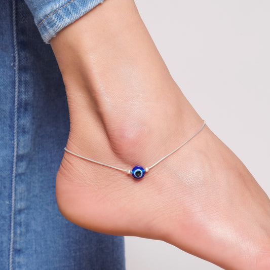 A silver anklet featuring a blue evil eye bead, worn on a person's ankle.